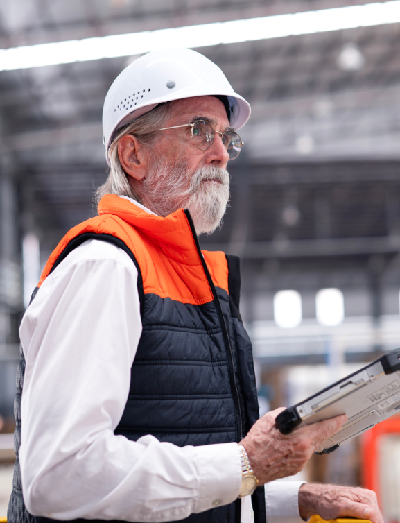 Older adult male in professional attire, wearing hard hat at construction site, holding mobile tablet.