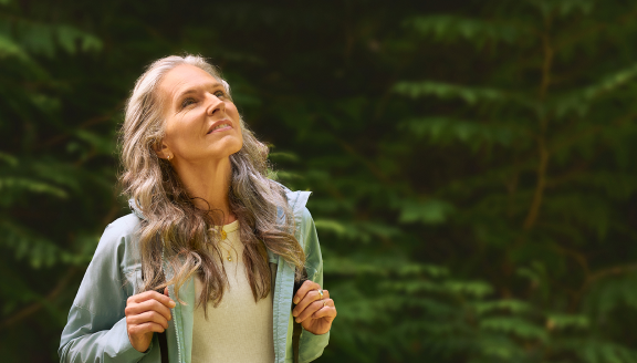 Adult woman in casual attire, with trees in background, looking at something above her in distance.