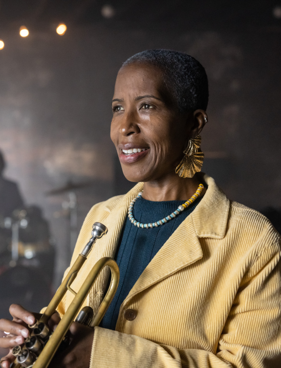 Adult woman holding trumpet indoor performance venue, with drummer in background.